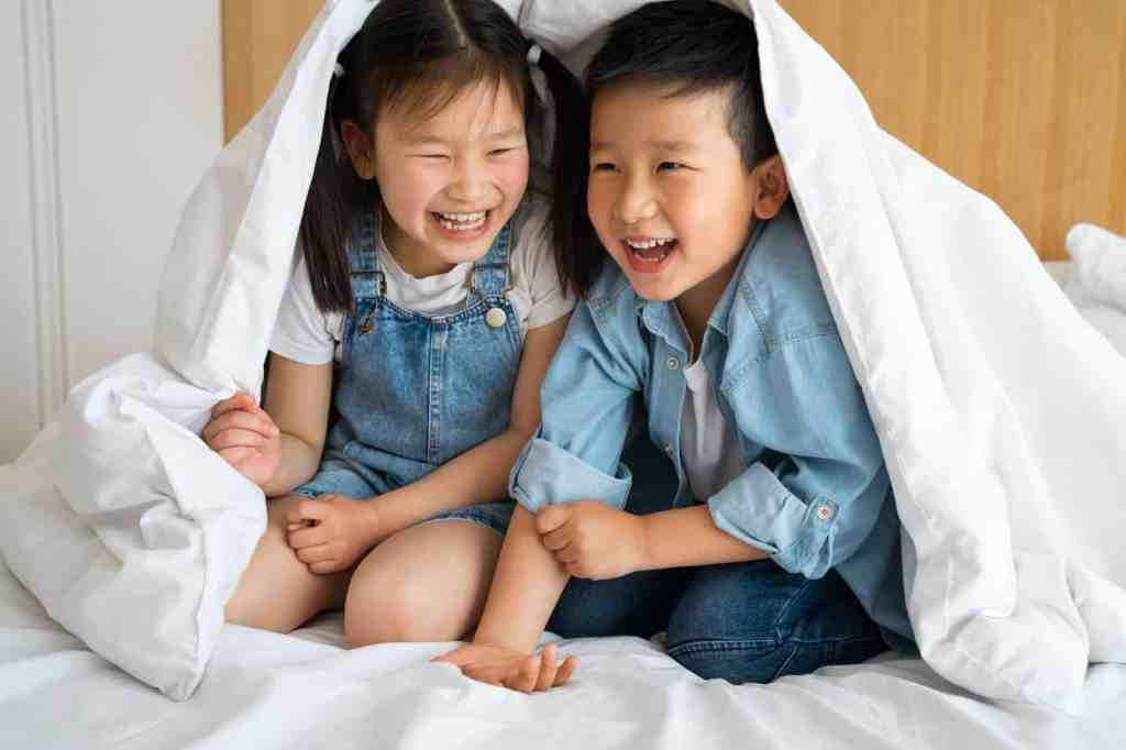 Two children laughing and playing together under a white blanket, sitting on a bed.