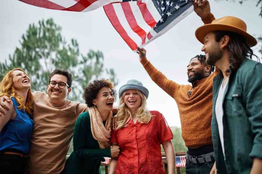 Diverse group of friends smiling and celebrating outdoors, holding and waving the American flag during a joyful gathering or patriotic event. Perfect for content related to travel, cultural diversity, or American festivals.
