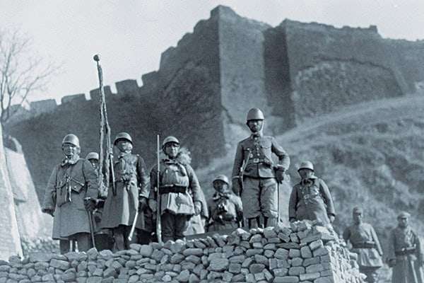 A group of soldiers in historical military uniforms posed in front of an ancient stone fortress, with a dramatic hillside in the background.