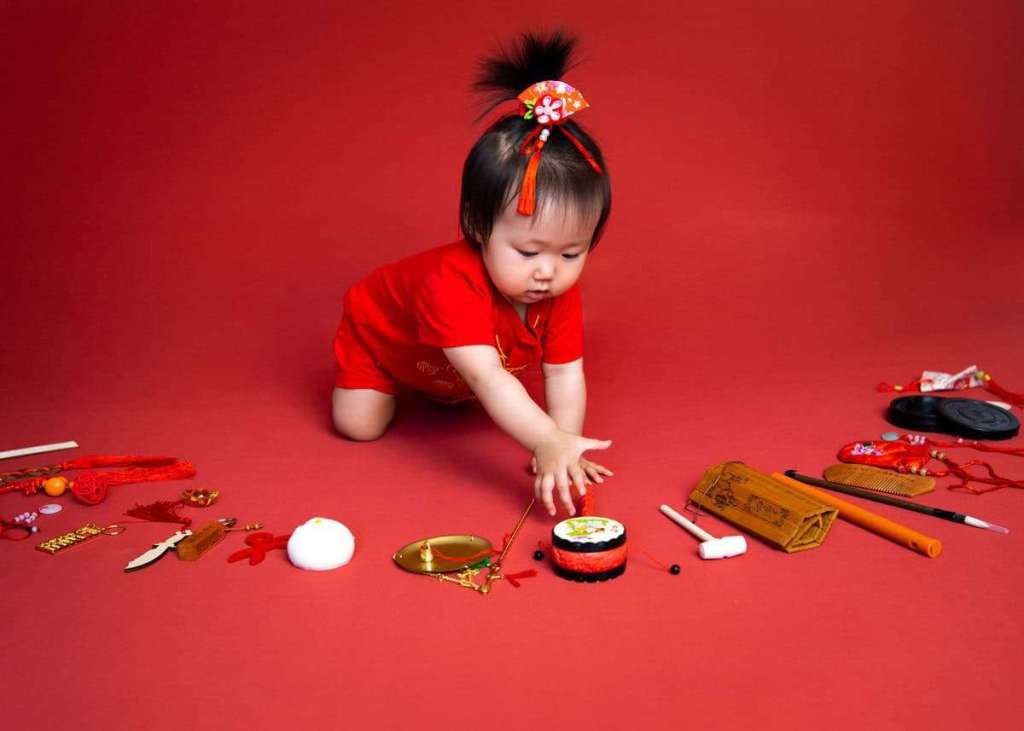 A young child dressed in red is playing on a red background, reaching for colorful decorations and toys, which include traditional symbols and items associated with festive celebrations.