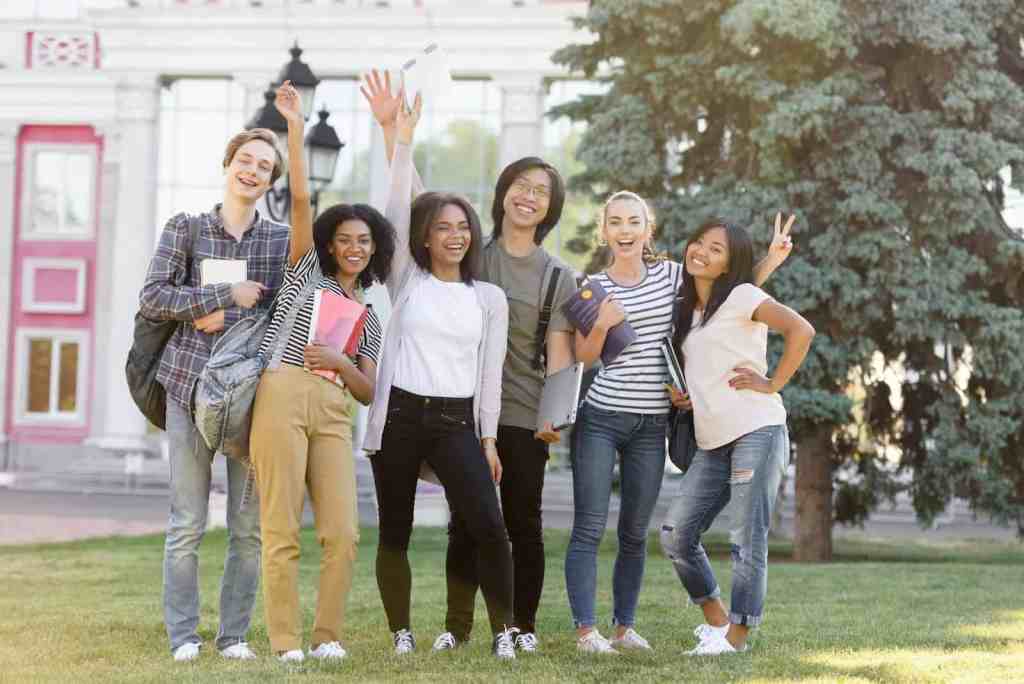 A diverse group of six young students standing outdoors, smiling and posing together. They are holding books and a laptop, with a backdrop of greenery and a building.