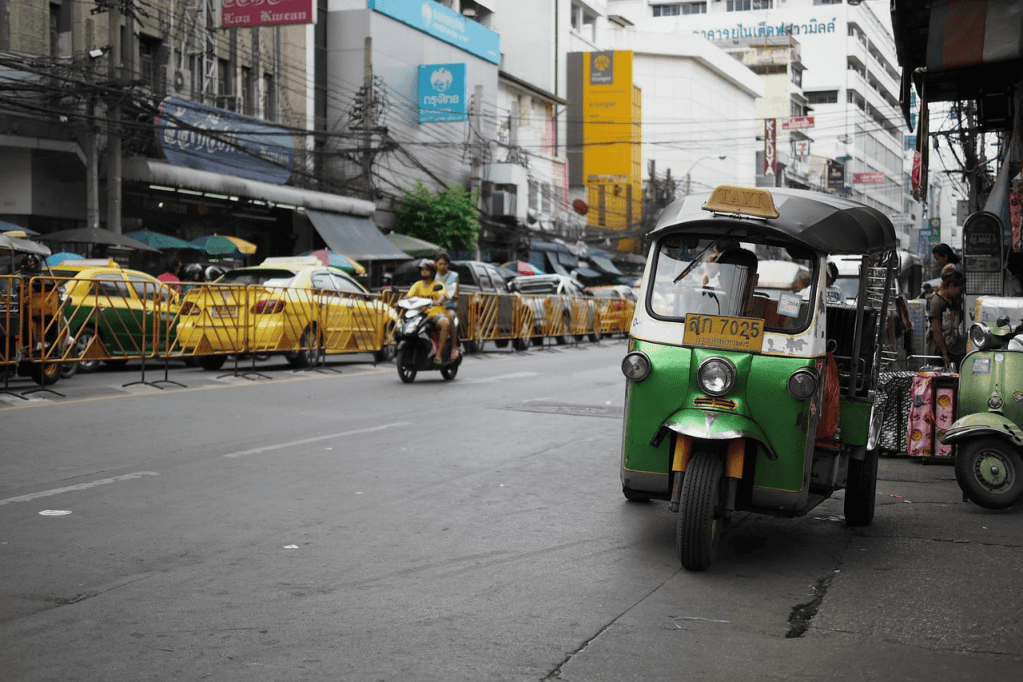 A green tuk-tuk parked on a busy street with yellow taxis lined up in the background. A motorcyclist is seen riding past, while barriers separate different areas of the road.