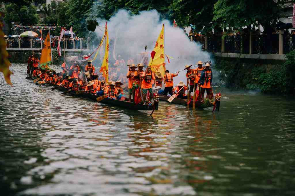 A colorful boat parade featuring participants wearing orange life jackets and traditional hats, while holding flags and paddling down a river filled with smoke.