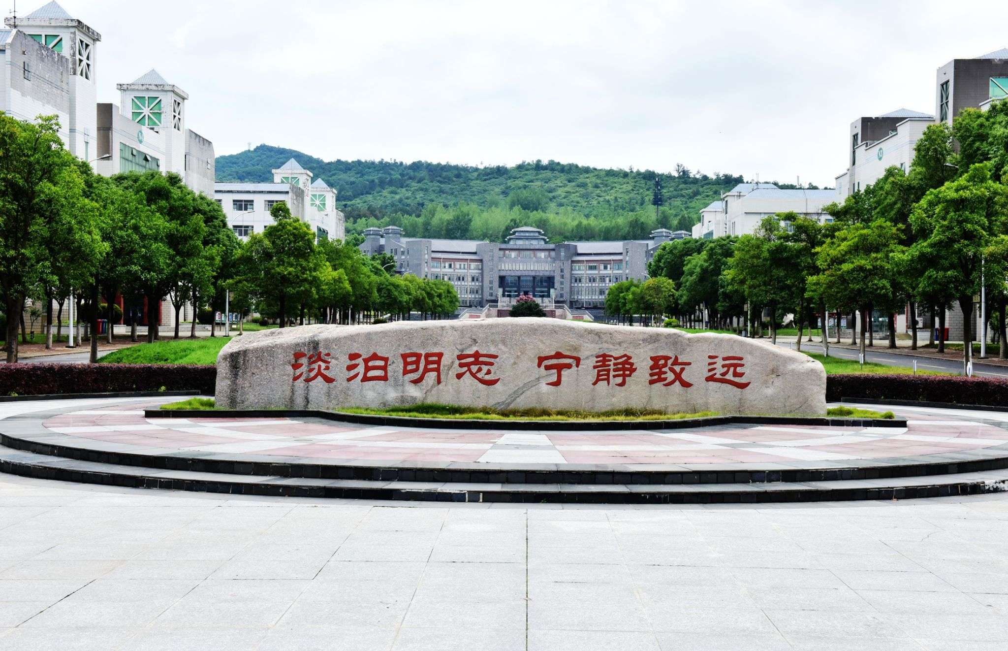 A stone monument with Chinese characters in front of a university campus, surrounded by trees and modern buildings, under a cloudy sky.