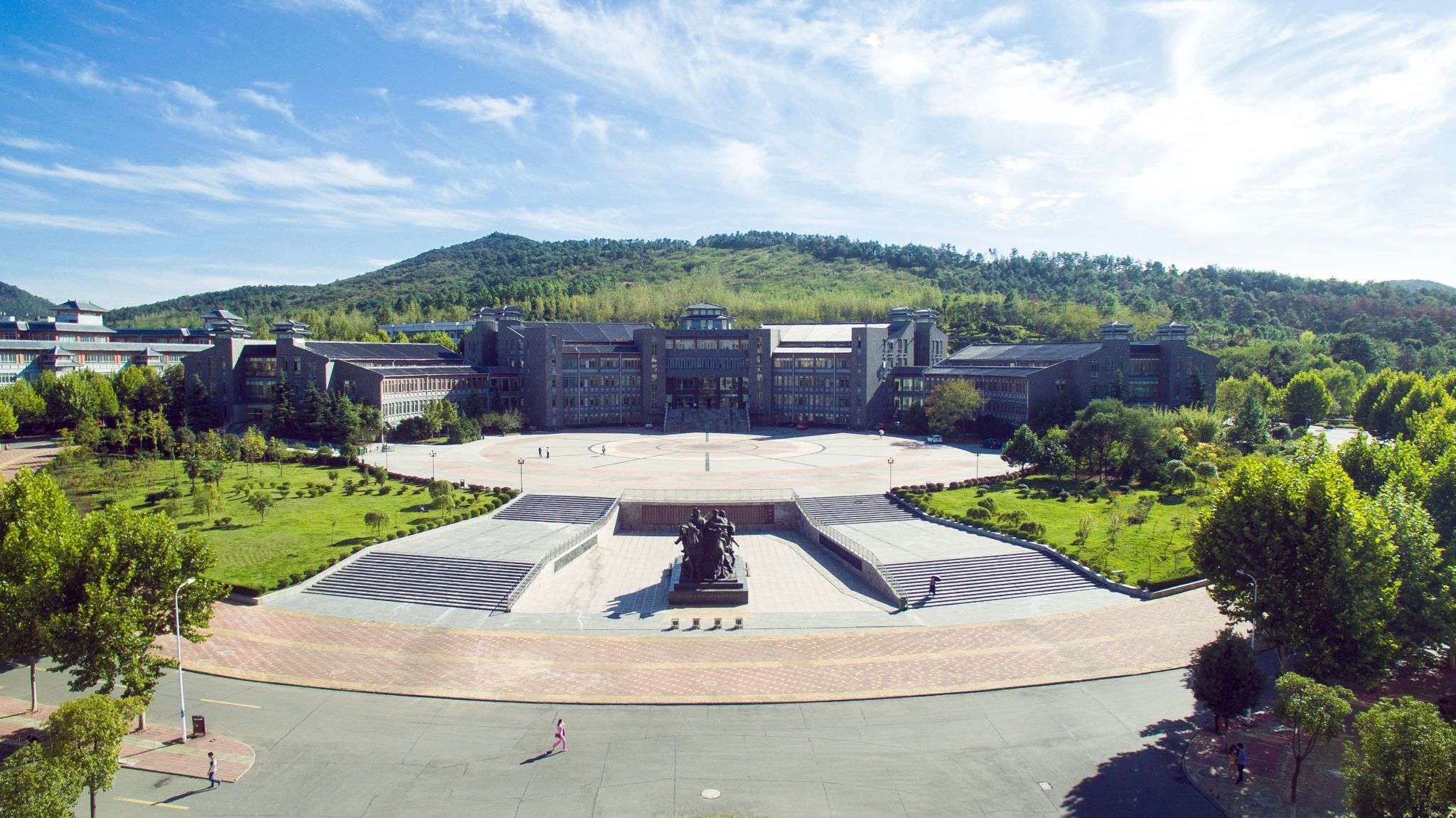 Aerial view of a modern educational building surrounded by landscaped gardens and trees, with a large courtyard in front and mountains in the background.