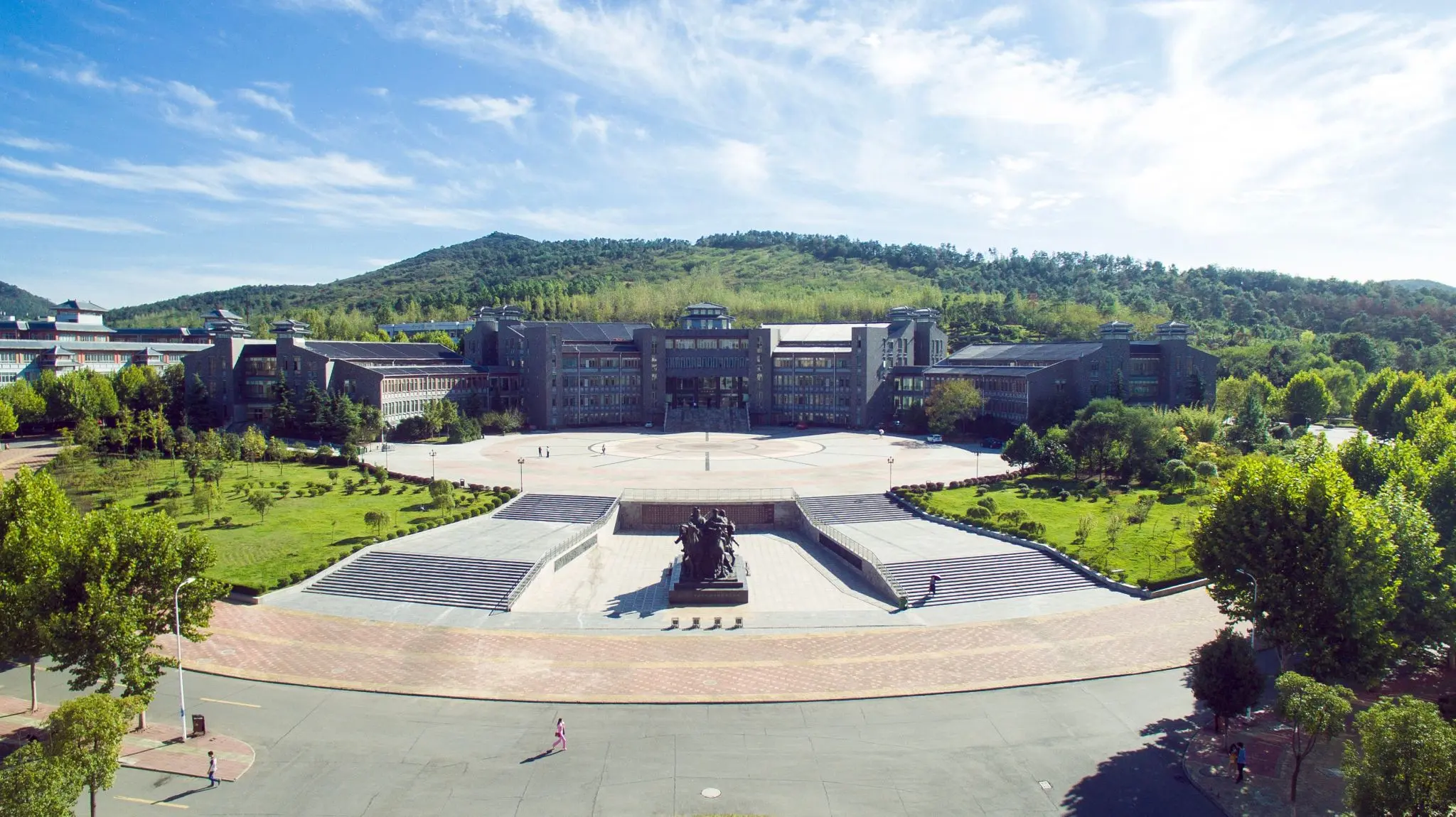 Aerial view of a modern educational building surrounded by landscaped gardens and trees, with a large courtyard in front and mountains in the background.