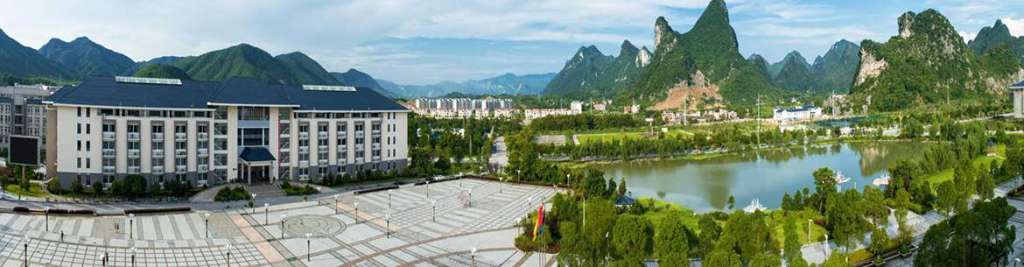 A panoramic view of a modern building next to a landscaped park and lake, surrounded by lush green mountains and clear blue sky.