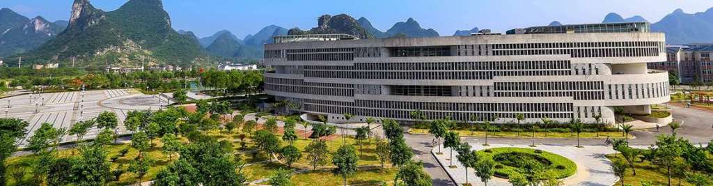 A modern building surrounded by lush greenery and trees, set against a backdrop of mountains and a clear blue sky.