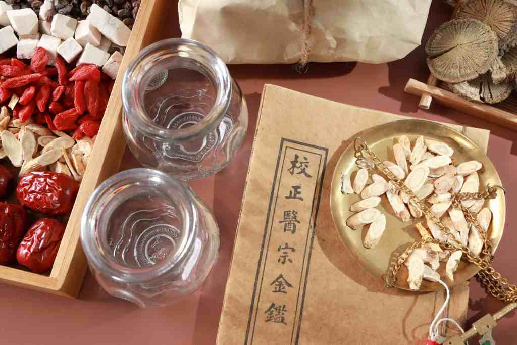 A collection of traditional herbal ingredients displayed in wooden trays, including goji berries, red dates, and various herbal slices. Two glass jars are positioned in the foreground alongside a gold weighing scale and a vintage book, suggesting a setup for herbal medicine preparation.