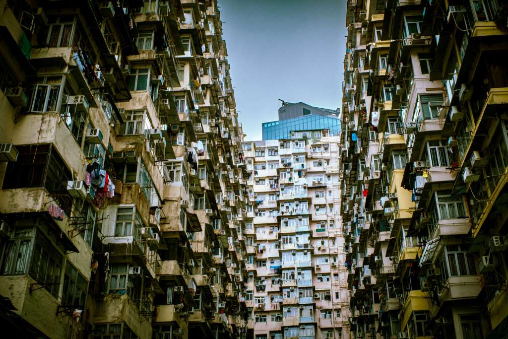 A narrow passage between two tall apartment buildings with numerous air conditioning units and clothes hanging outside the windows, under a clear sky.