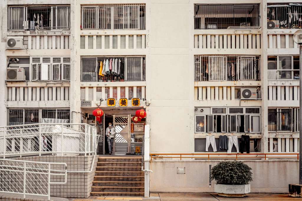 Exterior view of a residential building with a shop entrance, featuring red lanterns and a person standing by the door, and clothes hanging on balconies.