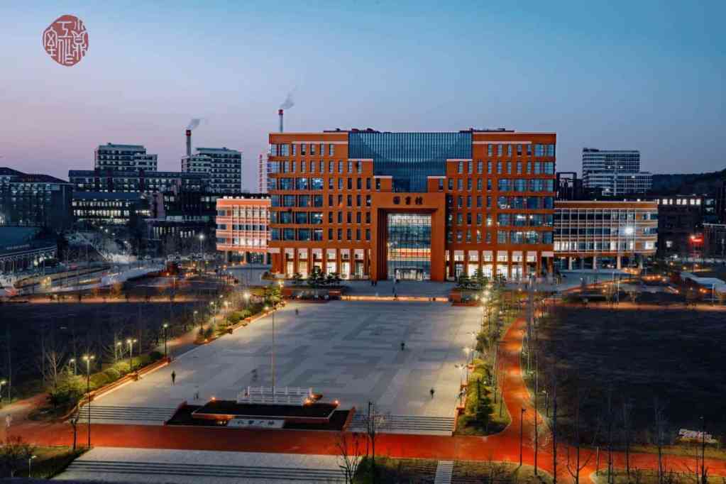 Aerial view of a modern red-brick building with large glass windows, surrounded by city buildings and a landscaped plaza at dusk.
