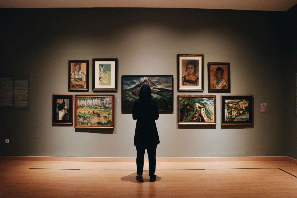 A person standing in an art gallery, observing a diverse collection of paintings displayed on a wall.