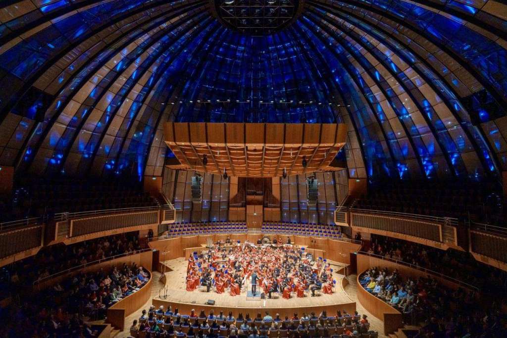 A large orchestra performing on stage in a modern concert hall with a striking glass ceiling illuminated by blue lights, and an audience watching from below.