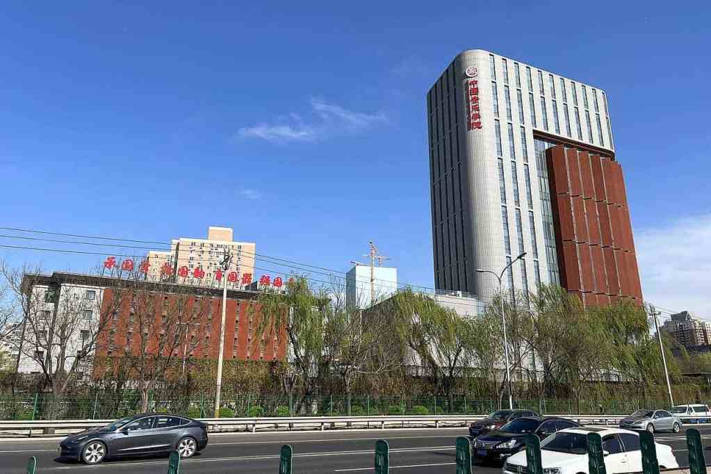 A modern building with a sleek design and a red panel on one side, set against a clear blue sky. The foreground features a road with cars and greenery along the sides.