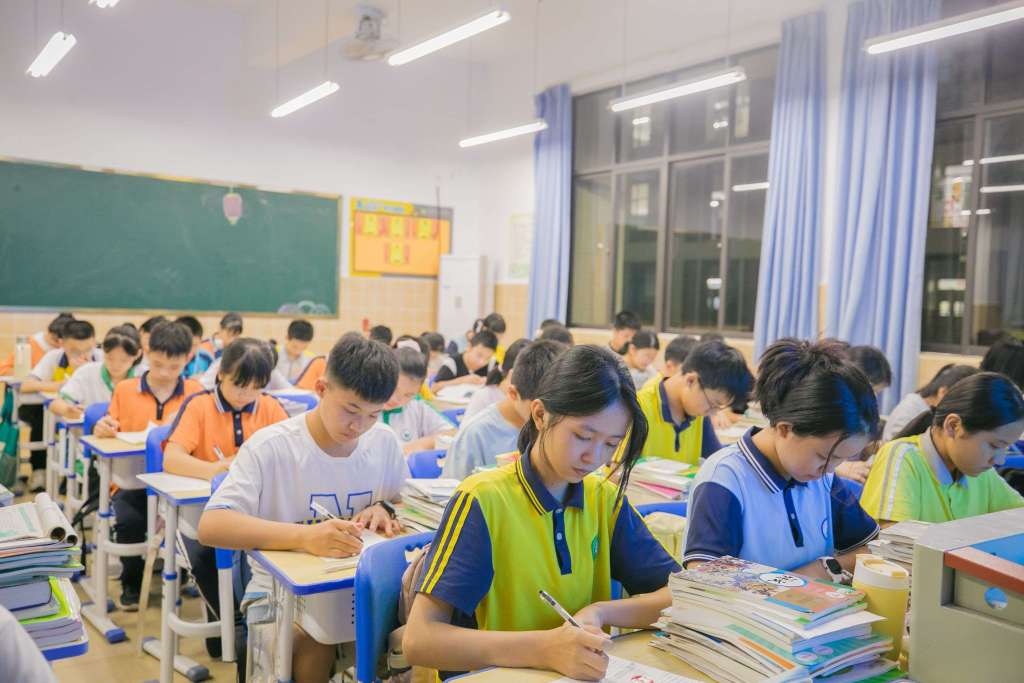 A classroom filled with students of various ages sitting at desks, focused on writing in their notebooks. The students are wearing colorful uniforms, and there are stacks of books on their desks. Bright classroom lighting and large windows are visible in the background.