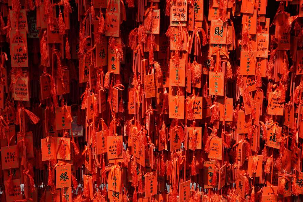 A wall covered with numerous bright orange tags, each decorated with black writings and tassels, often used for wishes or prayers in a cultural or religious setting.