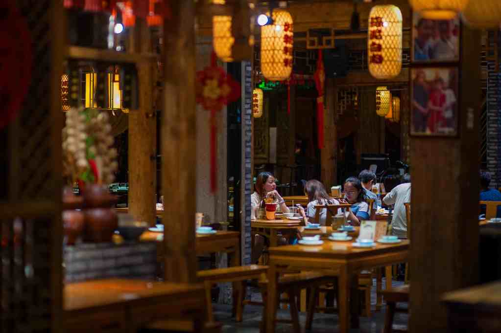Interior view of a restaurant with wooden tables and warm lighting, featuring patrons enjoying meals.
