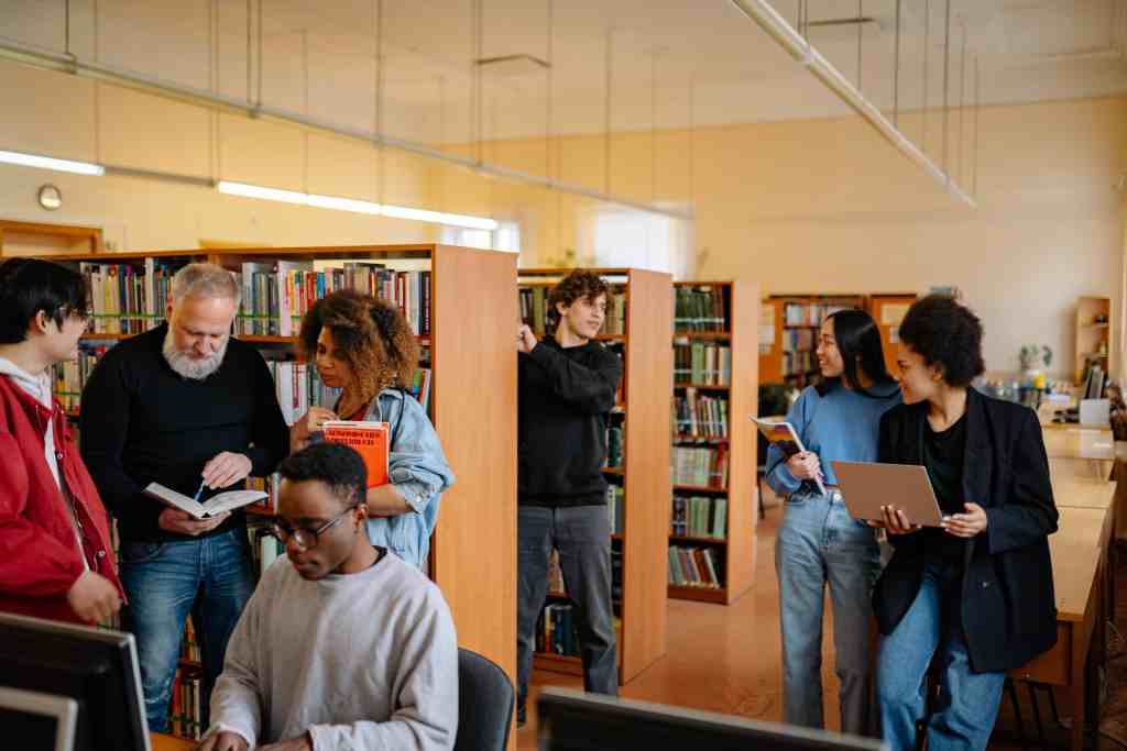 A diverse group of people interacting in a library setting, with some reading books and others discussing while seated at a computer. Bookshelves filled with books are visible in the background. international students study in China at a Double First-Class university campus