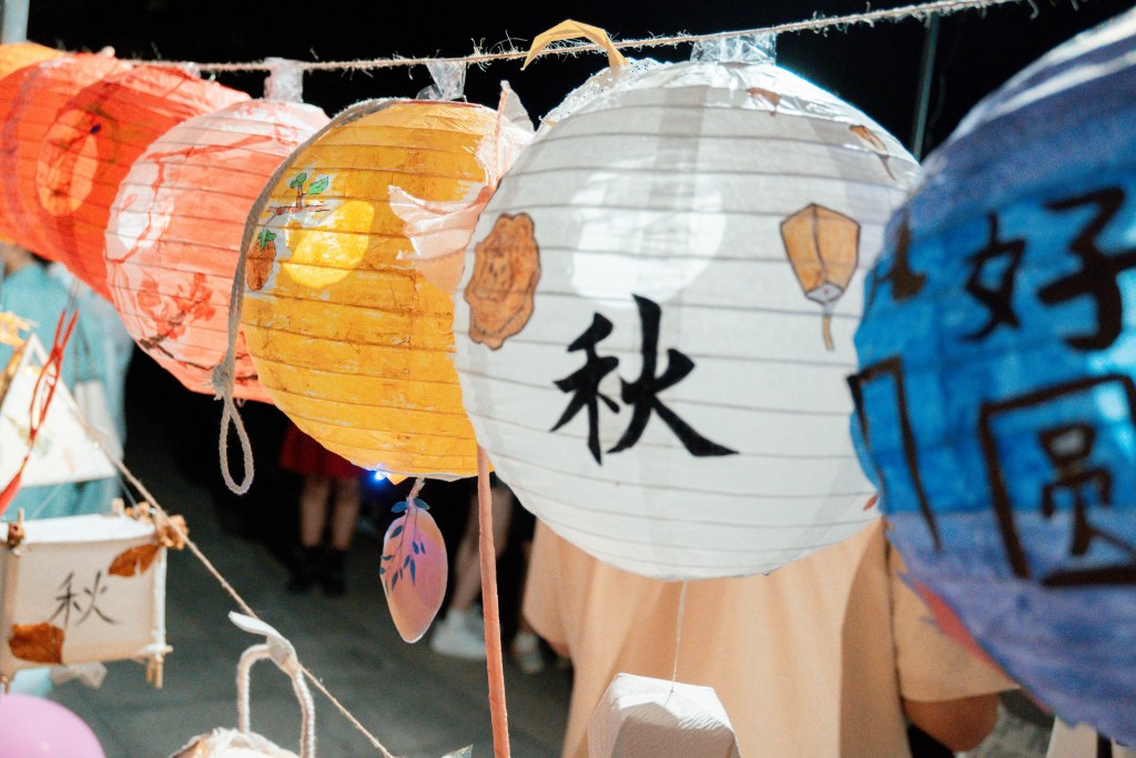 Colorful paper lanterns in various designs hanging on a string, with Chinese characters visible on some of them, set against a dimly lit background.