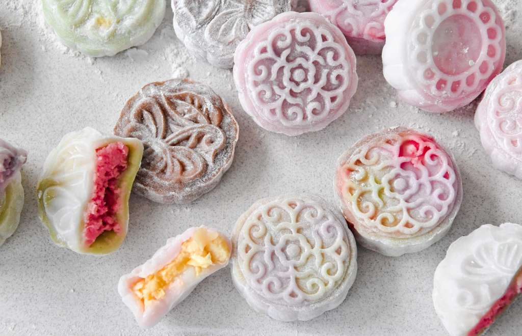 An assortment of decorative mochi desserts with colorful fillings, placed on a light surface. Some mochi are cut in half, revealing pink and yellow fillings, while others maintain their intricate designs.