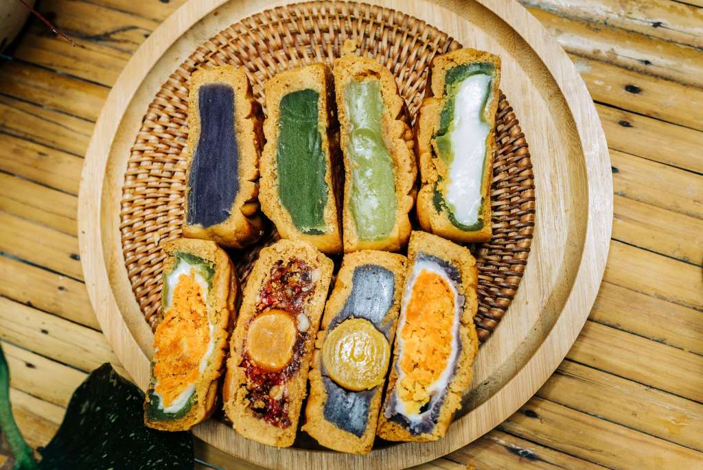 A wooden platter filled with colorful slices of traditional pastries, featuring layers of purple, green, orange, and white fillings with a variety of textures.