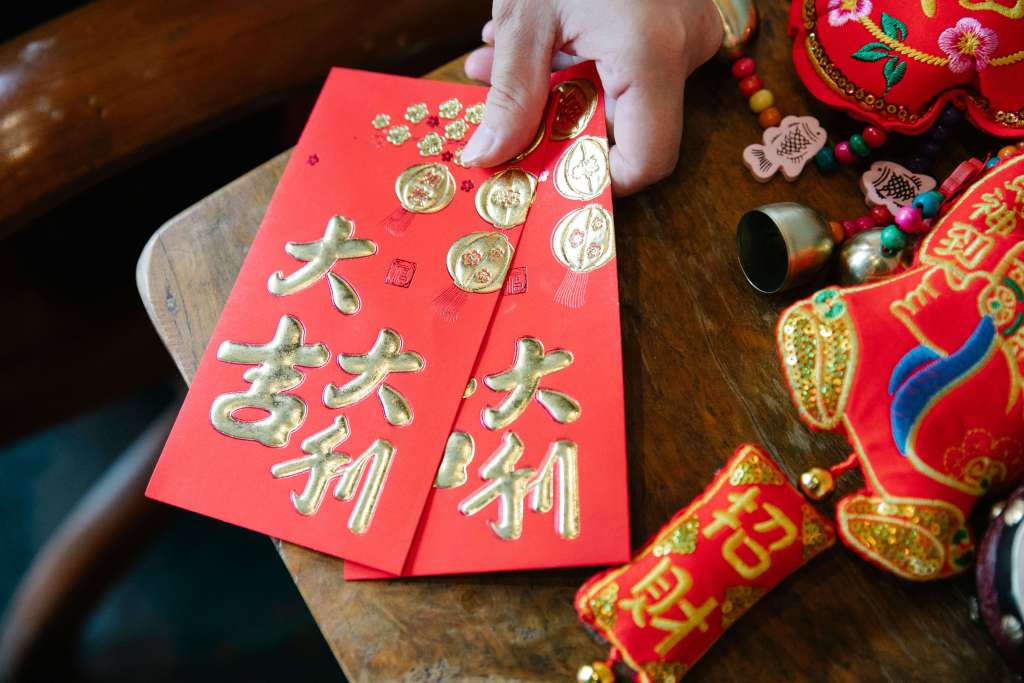 Two red envelopes with gold text and symbols placed on a wooden surface, accompanied by traditional Chinese ornaments and decorations.
